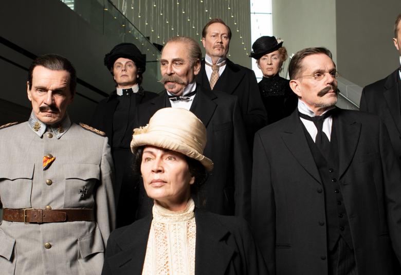 Kuvassa ovat näyttelijät portaikossa vuosisadan vaihteen tyylisissä vaatteissa ja olemuksessa.