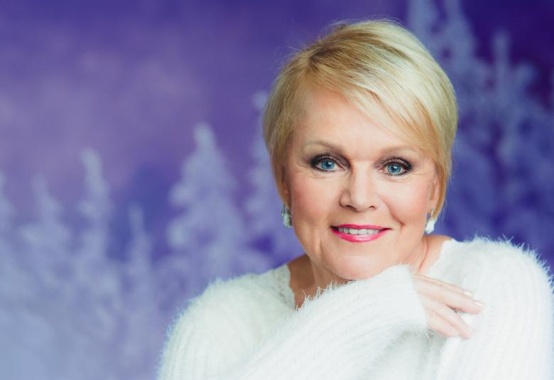 Kuvassa on Katri Helena puolivartalokuvassa ja hän on oikealla valkoisessa villapaidassa.  Tausta on heleän violetti ja siinä alaosassa on vaaleita kuusia.  