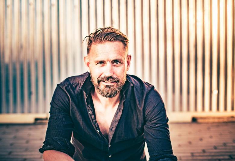 Kuvassa on Lauri Tähkä istumassa ulkona taustanaan pystysuoraa rimoista tehtyä seinää, joka on puun väristä.