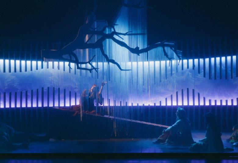 Kuvassa on tumman sinistä keskellä on vaalean sinistä valoa ja tummassa valossa on näyttelijöiden hahmoja istumassa.