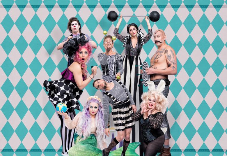 Kuvassa ovat näyttelijät rykelmänä seisomassa ja istumassa kirjavassa vaatetuksessa. Seinä on turkoosi-harmaan salmiakkikuvioinen.