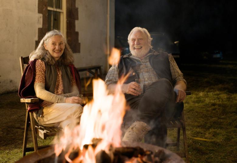 Kuvassa ovat Brid Brennan ja James Cosmo istumassa pimenneessä illassa nuotiolla sen takana istumassa. Vasemmalla on vaalean rakennuksen seinää.