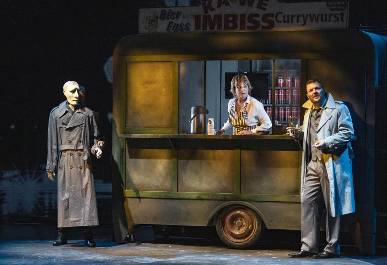 Kuvassa on keskellä ruskehtava vanhanaikainen kioskivaunu, jossa on ylhäällä kyltti Kawe Imbiss. vasemmalla on puukasvoinen mies  Jyrki MÄnttäri harmaassa tsenssissä ja oikealla  nojaa vaaleassa trenssissä ja puvusssa Tommi Raitolehto. Vaunussa on myyjänä Teija Auvinen.