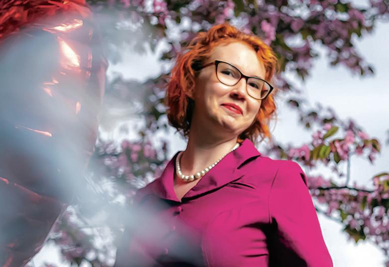 Kuvassa on Henna Karppinen-Kummunki puolivartalokuvassa ja violetissa mekossa. Taustalla on puu, jossa on violetin värisiä kukkia.