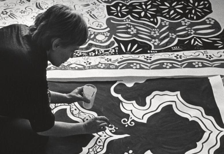 Kuvassa on Maija Isola lattialla kumartuneena maalaamaan kankaan kuvioita.  Kuva on musta-valkoinen.