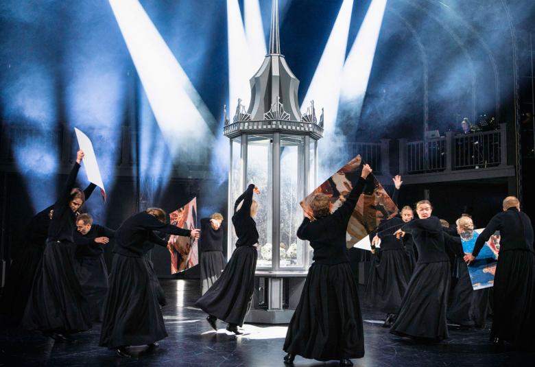 Kuvassa on esiintyjiä mustissa kaavuissa tanssimassa näyttämöllä ja yhdellä on käsissään tauluja ja keskellä on valkoinen lasitorni, johon suuntautuu valokartioita. 