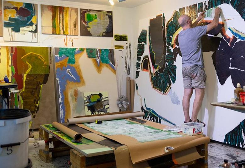Kuvassa on  taiteilija maalaamassa oikeassa reunassa maalaamassa isoa teosta seinälle.  Huoneessa on muita teoksia tekeillä ja keskellä työpöytä täynnä värejä sekä paperia.