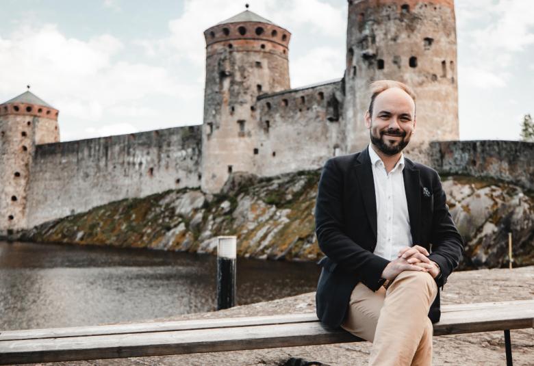 Kuvassa on Ville Matvejeff istumassa ulkona taustallaan Olavinlinna.  