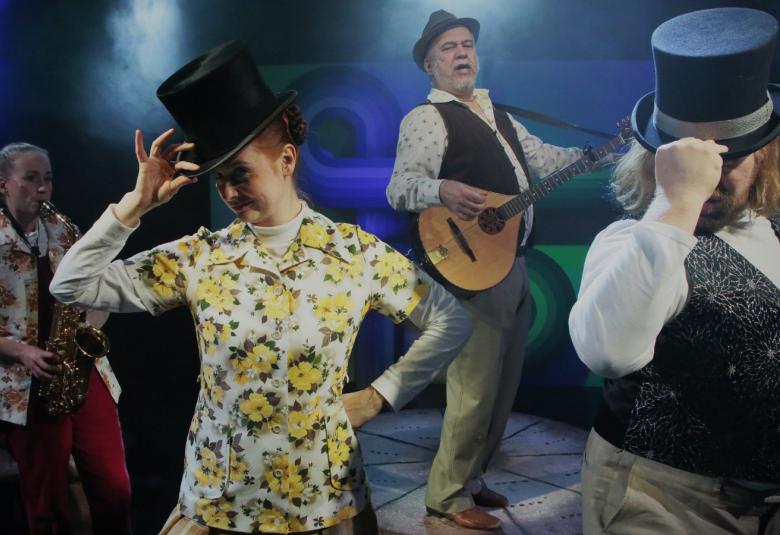 Kuvassa on neljä esiintyjää ja edessä on Saara Karjalainen pidellen toisella kädellä silinterihattua valko-vihreän kukallisessa puserossa. Taustalla Vera Veiskola ja takana keskellä Jari Rättyä soittamassa kitaraa hattu päässään ja mustassa liivissä. Edessä oikealla Janne Kinnunen silinteri päässään.