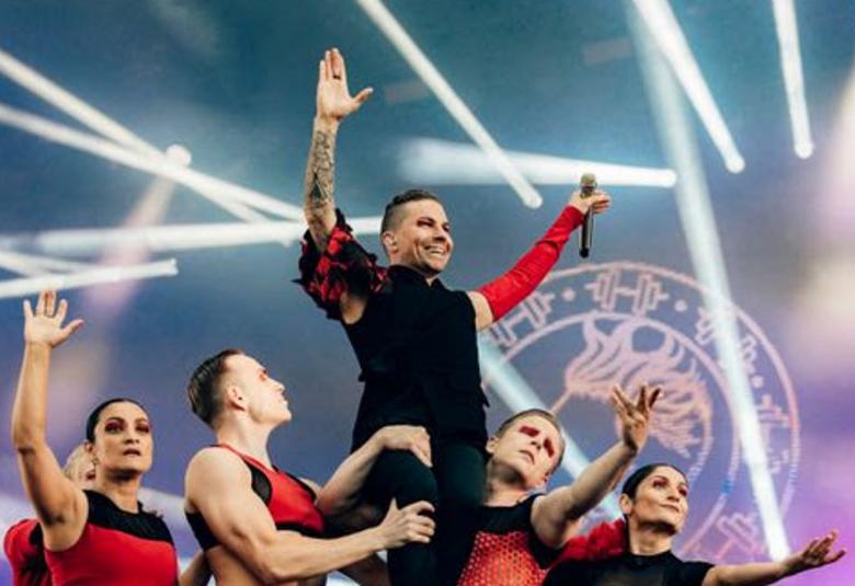 Kuvassa on Antti Tuisku lavalla alaviistosti kuvatusti.  Hän on keskellä kädet ylhäällä ja ympärillä alempana on viisi tanssijaa, joista näkyy päät. Tausta on taivaan sininen ja siinä on useampi valojuova.