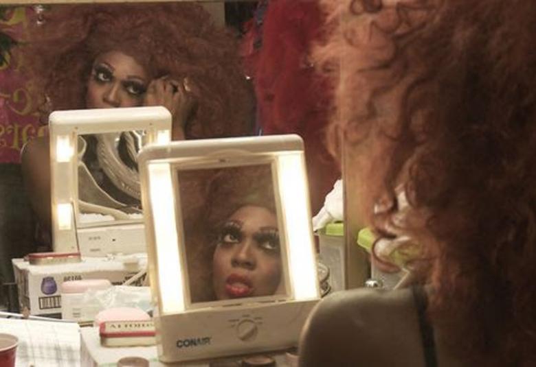 Kuvassa on tumma nainen meikkaamassa ja selin.  Hänen edessään on pieni iso peili ja sen takana on toinen pieni peili ja sen takana iso peili, josta näkyy kasvot.