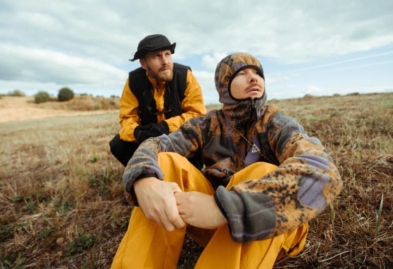 Kuvassa ovat Jare Brand ja Ville Galle istumassa ruuohikkoisessa rinteessa ja heillä tummaa ja keltaista päällään.