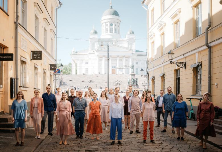 Kuvassa ovat kuorolaiset hajautuneina kadulle, jonka taustalla näkyy Helsingin tuomiokirkko. Vasemmalla ja oikealla olevat rakennukset ovat keltaisia.