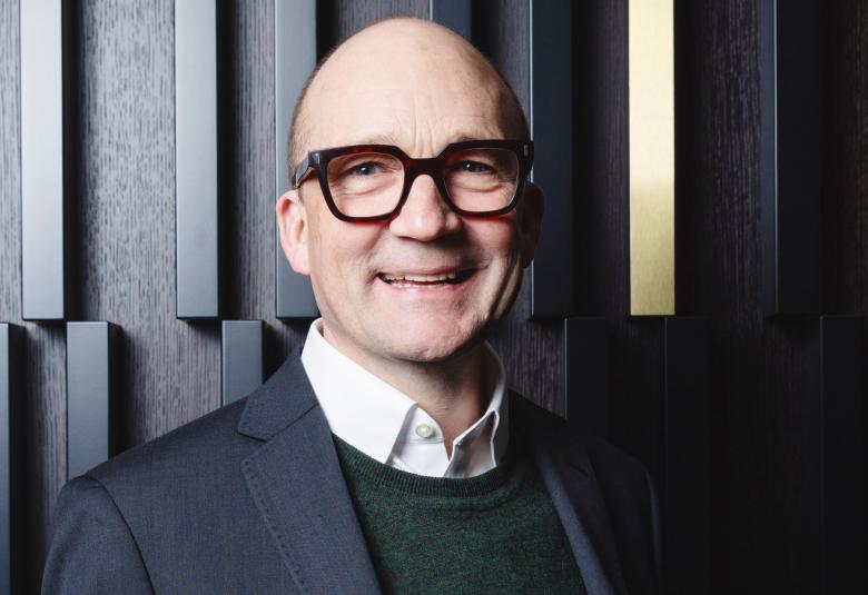 Kuvassa on Thomas de Mallet Burgess, josta on puolivartalokuva. Taustalla on mustaa seinää, jossa pieniä pystypalkkeja säännöllisin välein ja niistä on kaksi puun väristä.