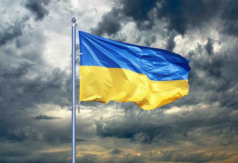 Kuvassa on Ukrainan sini-keltainen lippu liehumassa ja taustalla tummia pilviä.