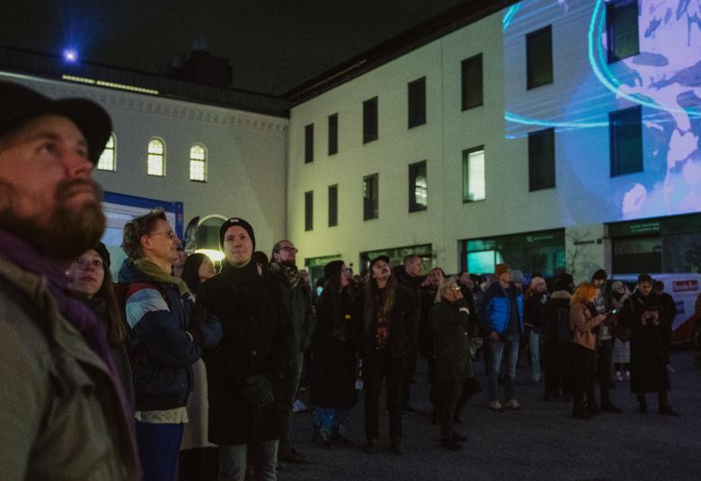 Kuvassa on kasvot eteenpäin ihmisiä katsomassa elokuvaesitystä sisäpihalla.  Kuvaa heijastuu rakennuksen vaalealle seinälle.