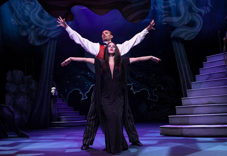 Kuvassa on kaksi näyttelijää, jotka ovat tumman näyttämön keskellä. Nainen on edessä kädet ovat nostettuina sivuille ja takana olevalla viistosti ylöspäin.