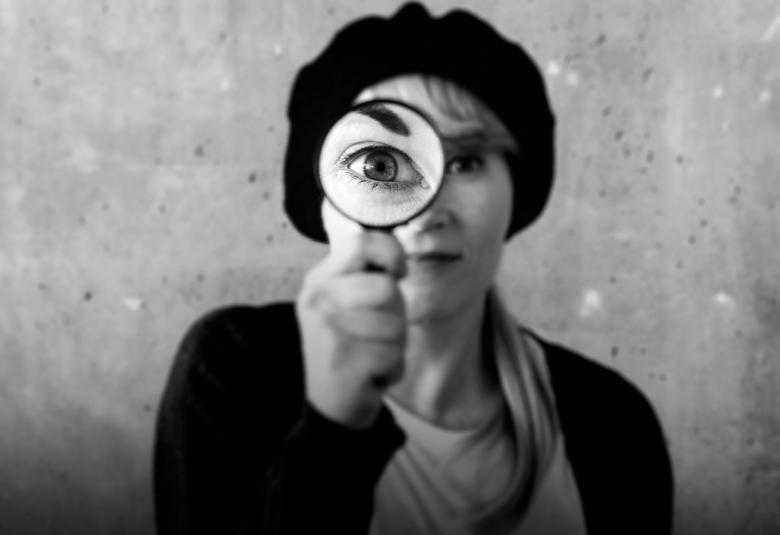 Kuvassa on nuori nainen joka näyttää silmänsä kohdalla suurennuslasia ja vasen silmä näyttää suuremmalta. Hänellä on baskeri päässään. Kuva on musta-valkoinen.