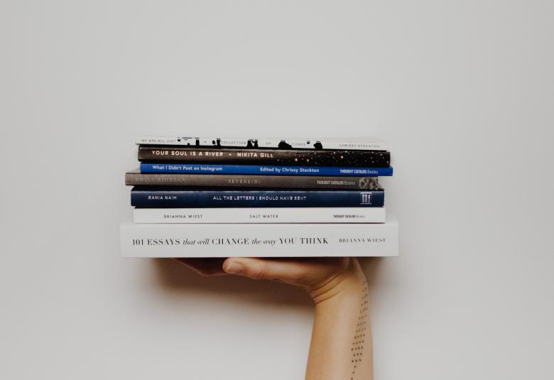 Kuvassa on käsi, joka pitelee kirjapinoa.  Kuvan tausta on harmaa.