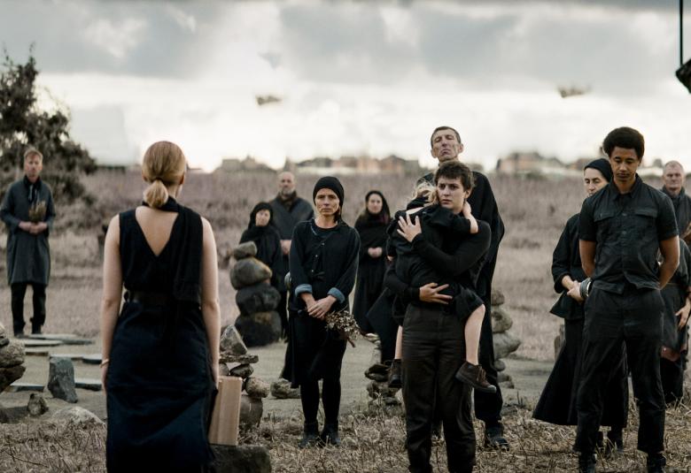 Kuvassa on harmaassa maisemamma mustiin pukeutuneita ihmisiä harvakseltaan ja selin on nuori nainen puhumassa heille. Maassa on kaksi matalaa kivistä koottua patsasta.