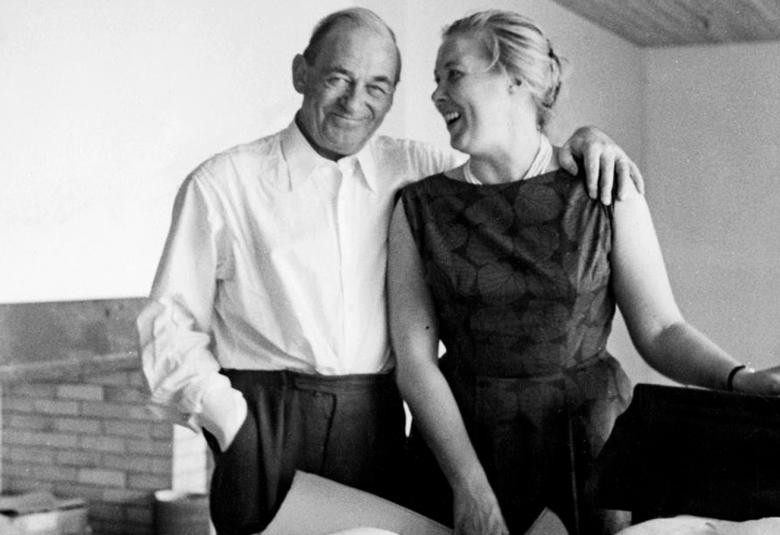 Kuvassa ovat arkkitehti Alvar Aalto ja hänen vieressään Elissa Aalto työhuoneellaan seisomassa työpöydän edessä ja Alvar Aalto pitää kättään Elissa Aallon olkapäällä. Kuva on musta-valkoinen.