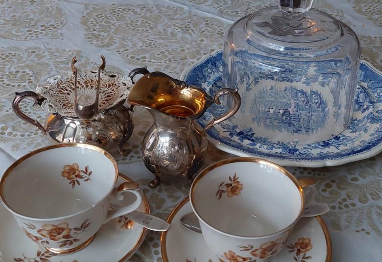 Kuvassa on kaksi Myrna-kuppia ja hopeinen sokerikko ja kermakko.  Sokerikon päällä on sokeriottimet.  Takana on lasikupu.