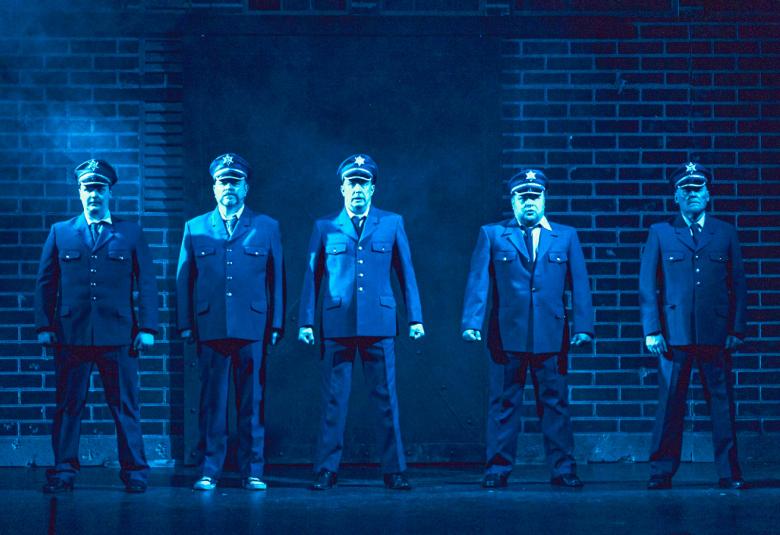 Kuvassa on viisi miestä sinisissä virkapuvuissa ja koppalakeissa.  Taustalla on tiiliseinää.  Kuvan sävy on sinertävä.