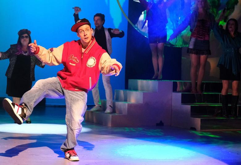 Kuvassa on oikealla portaikko ja siinä himmeästi näkyy näyttelijöitä.  Keskellä on nuori mies, joka tekee vinohyppyä ja hänellä on päällää puna-harmaa pusakka.