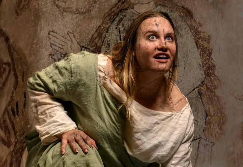 Kuvassa on Aurora Manninen kumartuneena ja hänellä on päällään vaalean vihreä karkeatekoinen mekko. Taustalla on seinämaalauksia.