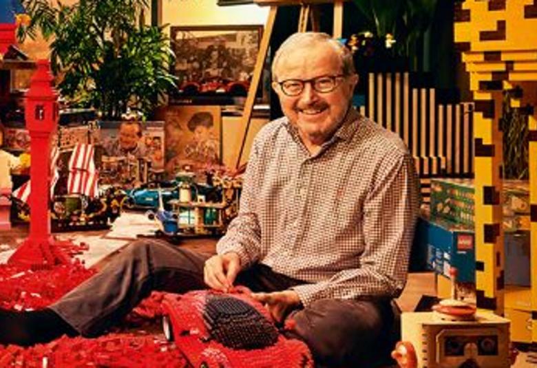 Kuvassa on Ole Kirk Christiansenisumassa legojen keskellä.  Vieressä on korkea kelta-ruskea legotorni.