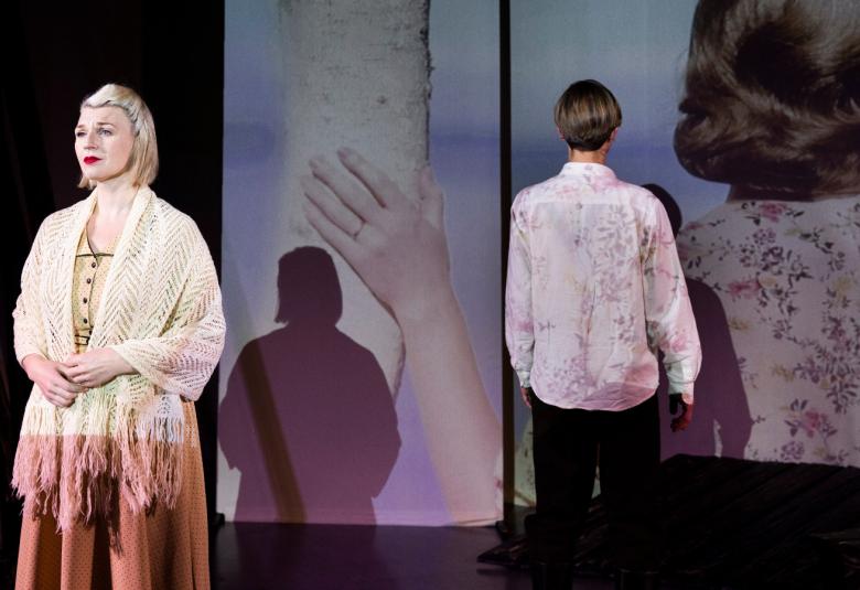 Kuvassa on Regina Linnavuo valkoinen villahuivi harteillaan seisomassa yleisöön päin. Takana seisoo selin nuori mies. Heidän takanaan on heijastettu kuva, jossa on nuoren naisen pää ja selkää takaa päin. Hän pitää kättään koivun rungolla. 