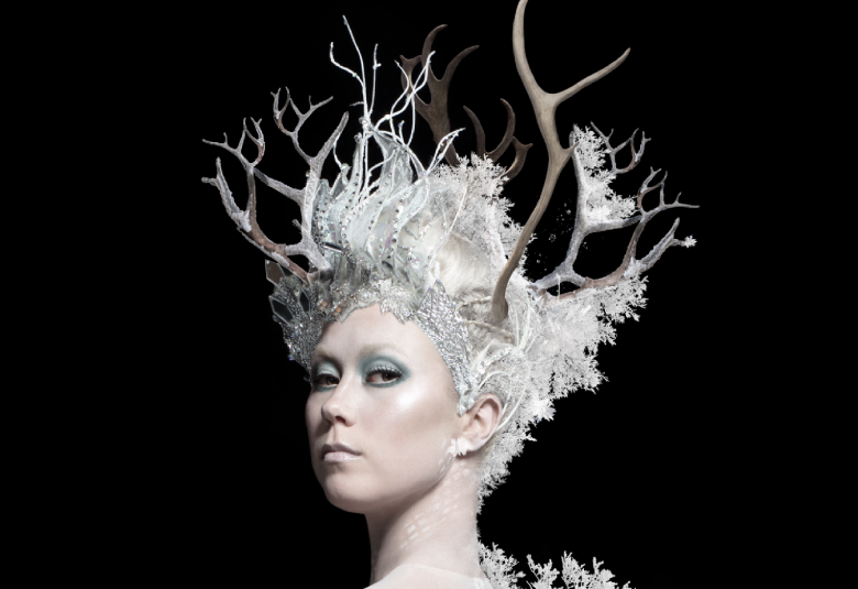 Kuvassa on tanssijan kasvot ja hänellä on päässään hiukset ylös nostettuna ja oksainen, valkoinen huurteinen päähine. Tausta on musta.