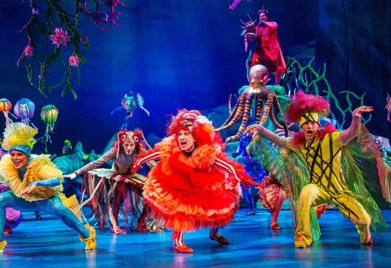 Kuvassa on värikkäissä vaatteissa olevia merenalaisia eläviä esitteviä tanssijoita näyttämöllä.