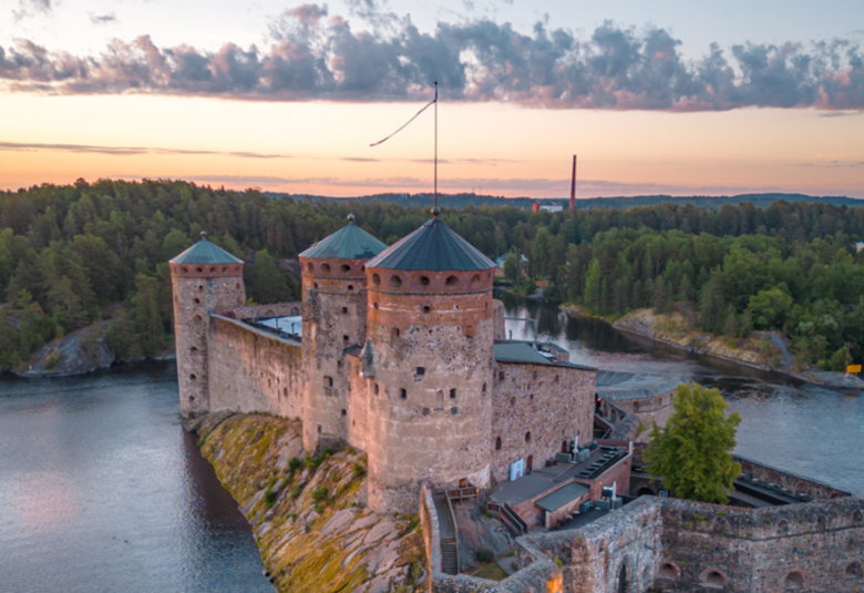 Kuvassa on Olavinlinna kuvattuna ilmasta ja yläosassa on sinertävän vaalean ruskeata pilvimuodostelmaa.