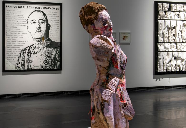 Kuvassa on etualalla värikäs naisfiguuriveistos, jossa ei ole jalkoja.  Taustalla näkyy tauluja.