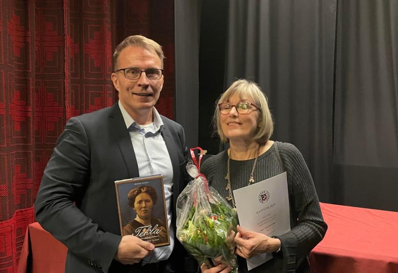 Kuvassa ovat Jukka Kopra kädessään kirja Tekla Hultinista ja Helena Kujala kädessään kukkakimppu ja kunniakirja. Kuva on puolivartalokuva ja taustalla on tumman punaista ja tumman harmaata verhoa.