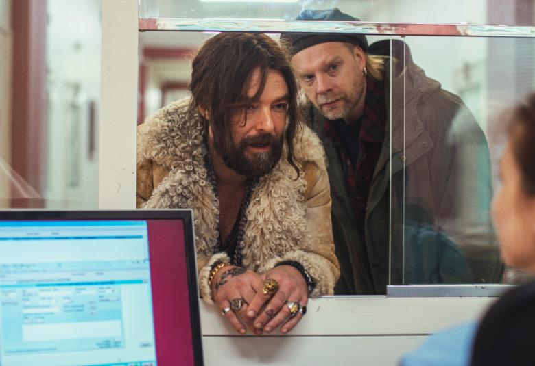 Kuvassa on Mikko Leppilampi palvelutiskin lasin takana keskustelemassa virkailijan kanssa.  Leppilammella on roikkuvat hiukset ja partaa.  Takaa näkyy Ville Myllyrinne kurkkimassa olkapään yli.