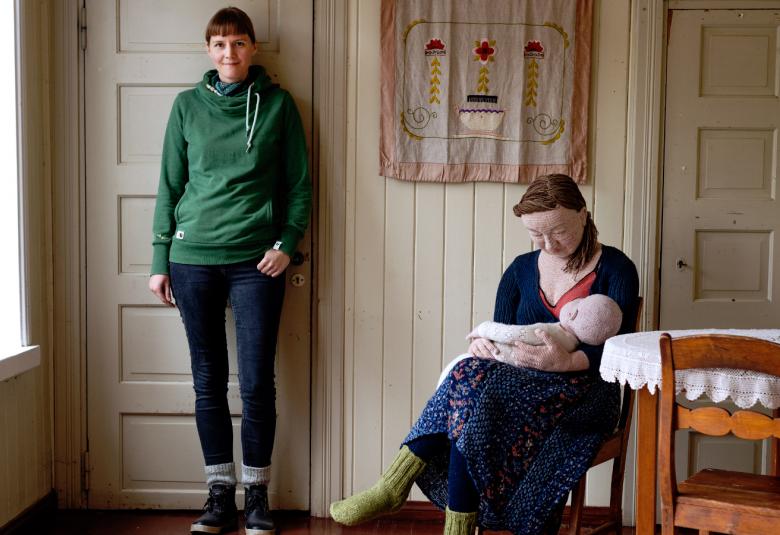 Kuvassa on Liisa Hietanen seisomassa huoneen seinään nojaten ja vierellä on tuolissa istumassa nainen, jolla on vauva sylissään. Nämä ovat virkattuja.