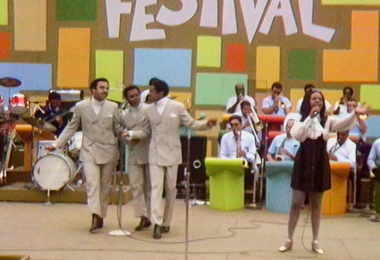 Kuvassa on kuva festivaalilavalta, jossa on taustalla iso bändi ja taustaseinällä on eri värisiä suorakaiteen muotoisia kuvioita. Edessä on oikealla vaaleissa puvuissa olevia mustia mieslaulajia ja oikealla musta naislaulaja.