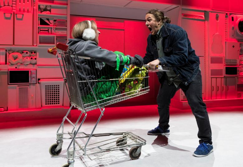 Kuvassa on mies työntämässä ostoskärryjä ja lastina on nainen.  Kuvan tausta on punainen.