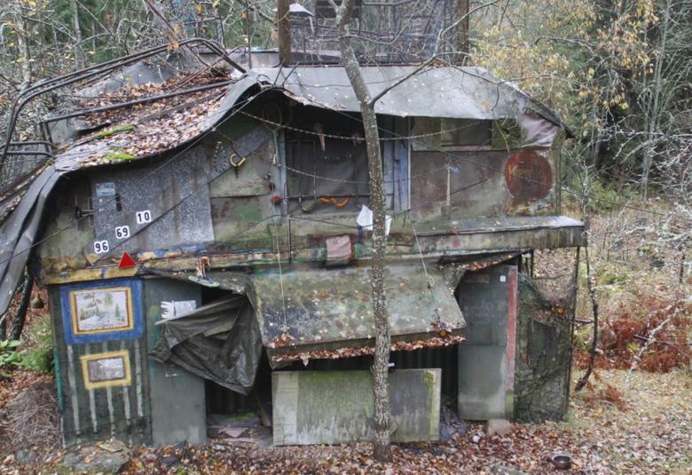 Kuvassa on metsässä harmaa lainemainen harmaa Villa Mehu, jossa on numeroita seinässä ja oksia ym. Se on naamioitu maastoon.