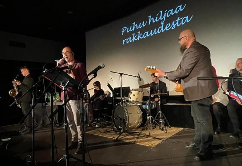 Kuvassa 3 on taustalla kappaleen nimi Puhu hiljaa rakkaudesta ja edessä laulaa mies kitaran kanssa pikkutakki päällä.