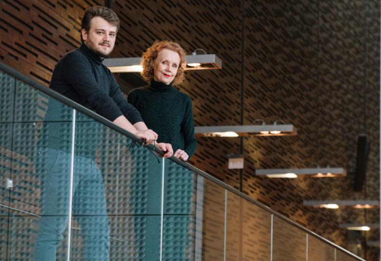 Kuvassa ovat Aleksi Barrière ja Kaija Saariaho kuvattuna alhaalta päin nojaamassa kaiteeseen, joka on harmaata paksua verkkoa. Taustalla on ruskeaa seinää ja kattoa.