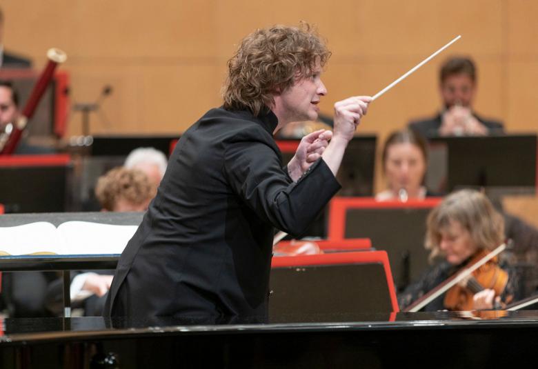 Kuvassa on Santtu-Matias Rouvali johtamassa Lontoon filharmonista orkesteria, jonka soittajia näkyy hänen takanaan.  Rouvali on kuvassa vinottain.