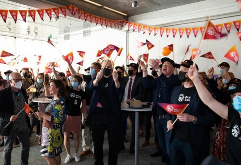 Kuvassa on Oulun kulttuuripääkaupunki-päätöksen juhlimista sisätilassa teltassa.  Ylhäällä on oranssia viirinauhaa ja yleisö heiluttaa oransseja viirejä.