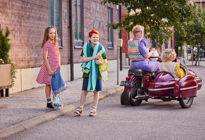 Kuvassa ovat vasemmalla seisomassa kadulla Nelli Nuudelipää sini-valkoraidallisessa mekossa ja Risto Räppääjä shortseissa ja t-paidassa.  Vieressä ovat moottoripyörän päällä Pakasta-Elvi ja punaisessa sivuvaunussa Rauha Räppääjä.