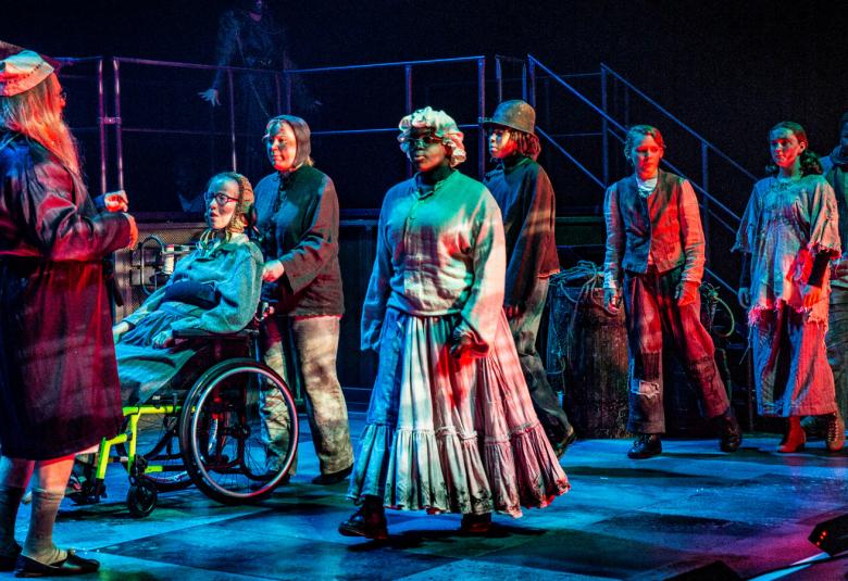 Kuvassa on näyttämöllä Eppu Normaali Saiturina selin vinottain ja hänen edessään lapsia jonomaisessa muodostelmassa pukeutuneina hieman resuisiin vaatteisiin.  Valaistus on tumma.