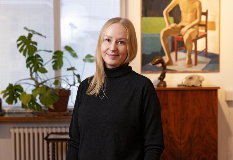 Kuvassa on Sanna Lipponen mustassa mekossa taustallaan ikkuna, jonka vasemmalla puolella on viherkasvi.  Oikealla on seinällä maalaus alla ruskea kaappi.