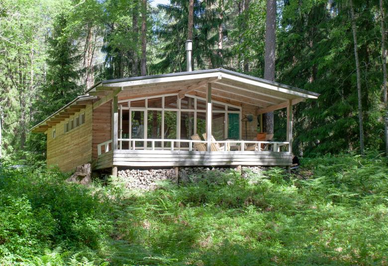 Kuvassa on matala, vaalea kesämökki, joka on vihreän lehtipuustoisessa maastossa.