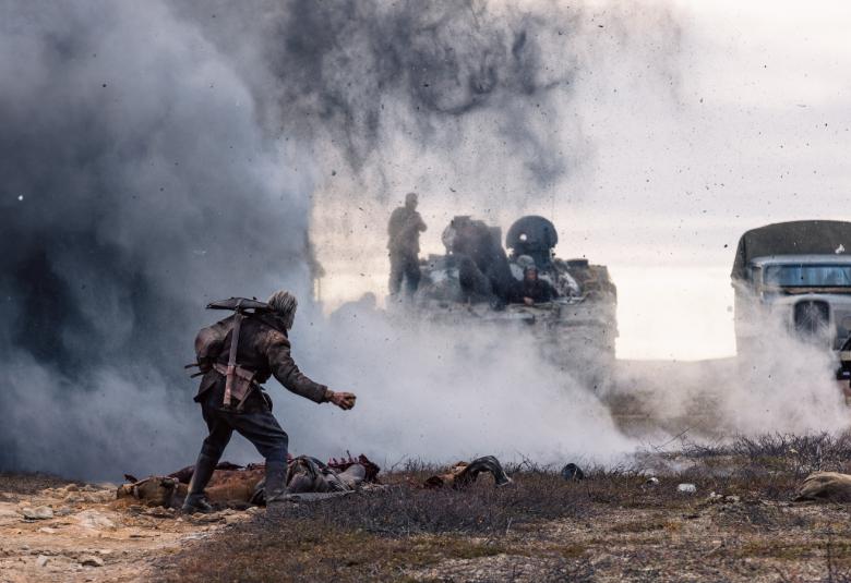 Kuvassa on etualalla kumarassa kulkeva Jorma Tommila ja hänen edessään on savua ja panssarivaunu miehistöineen. Oikealla on miehistönkuljetusauto.